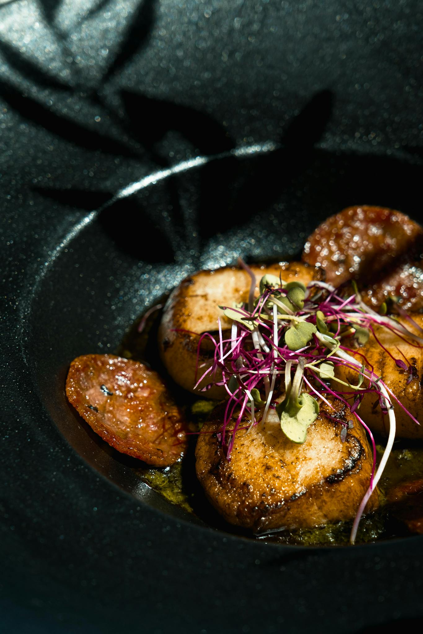 Seared scallops paired with chorizo and microgreens served on stylish dark plate.